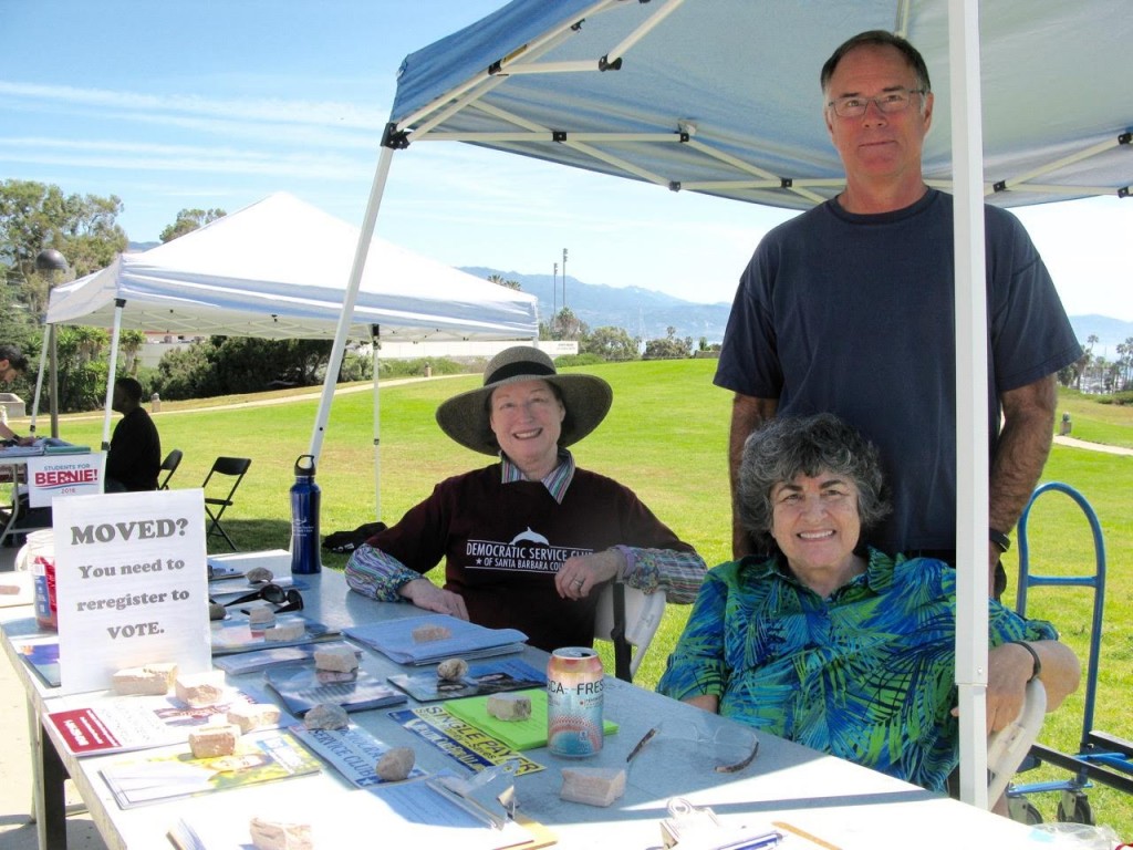 three people with registration materials on a desk in front of them and a green lawn behind